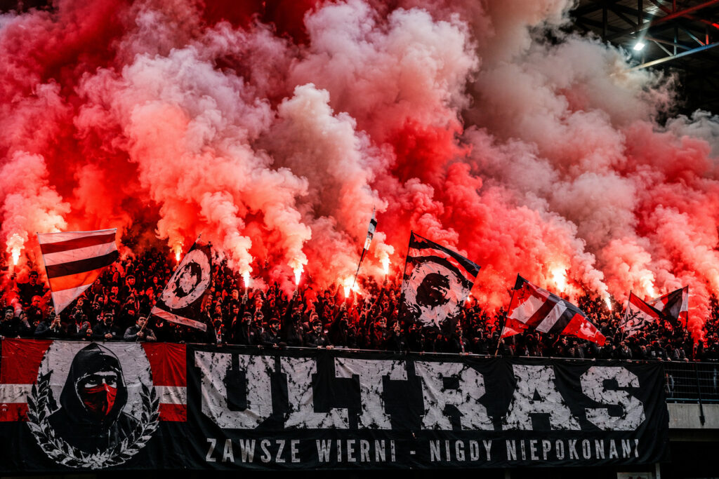 Kibice prezentujący oprawę meczową z wykorzystaniem czerwonych i białych świec dymnych na stadionie